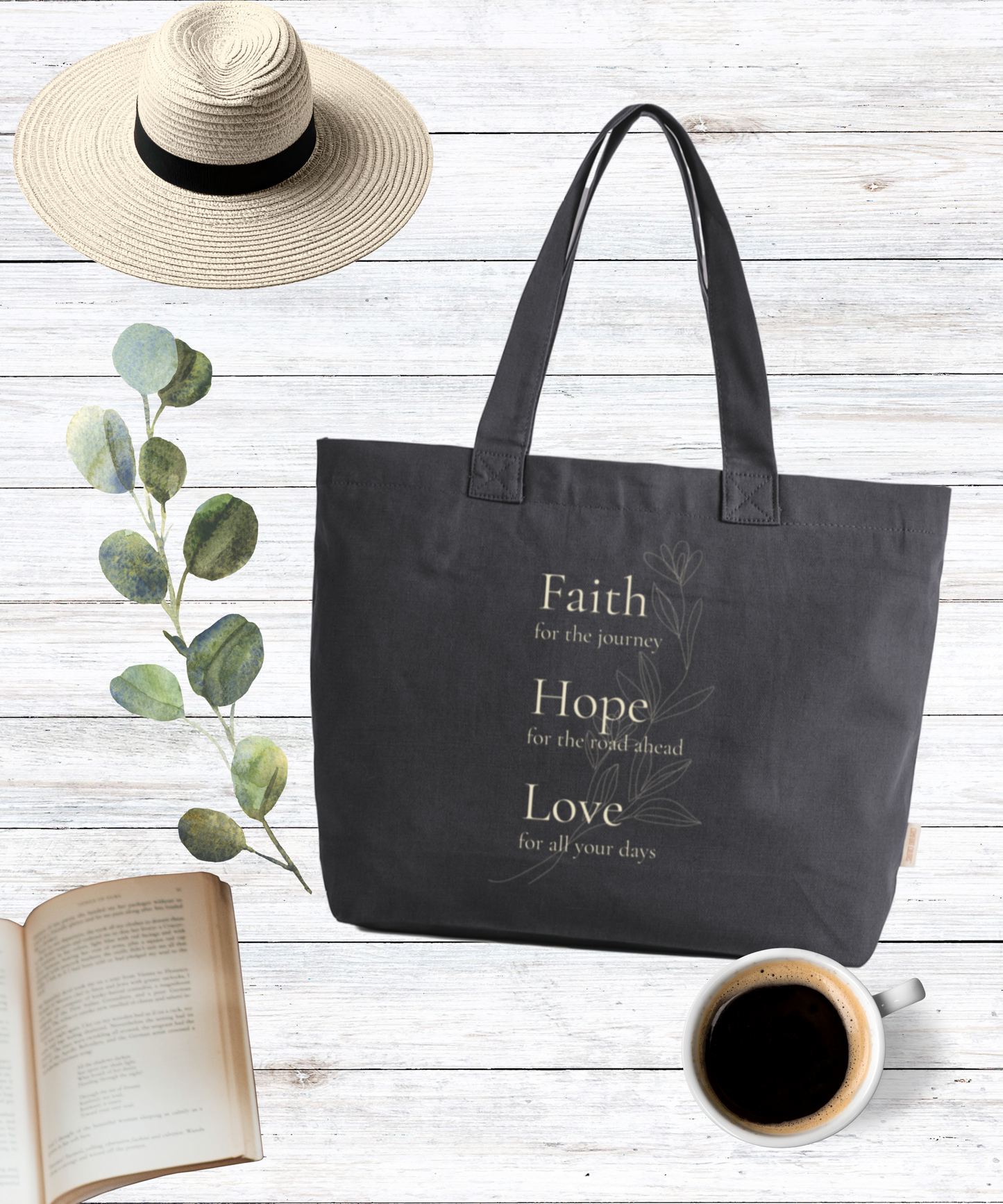 Graphite Comfort Colors zip tote bag with cream Faith, Hope, Love floral design, styled with eucalyptus, coffee, book, and straw hat on a rustic white wood background.