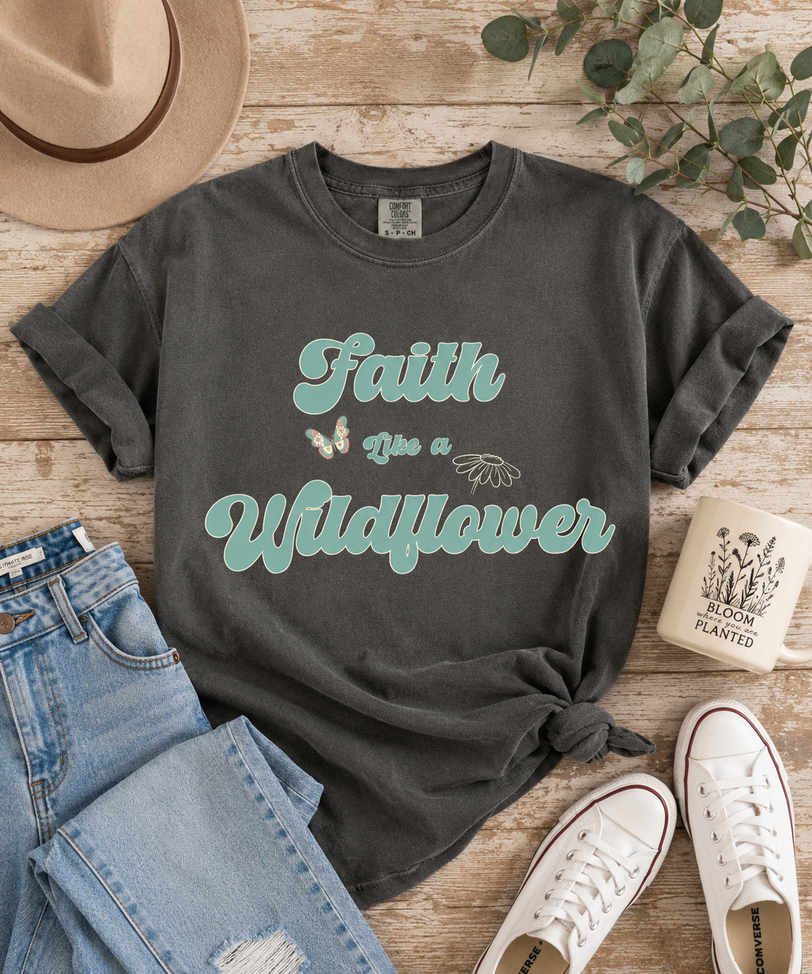 Flat lay of a Comfort Colors pepper t-shirt featuring the Faith Like a Wildflower design in soft teal and cream with butterfly and botanical accents. Styled with light denim jeans, white sneakers, greenery, and a rustic background for a vintage-inspired lifestyle look.