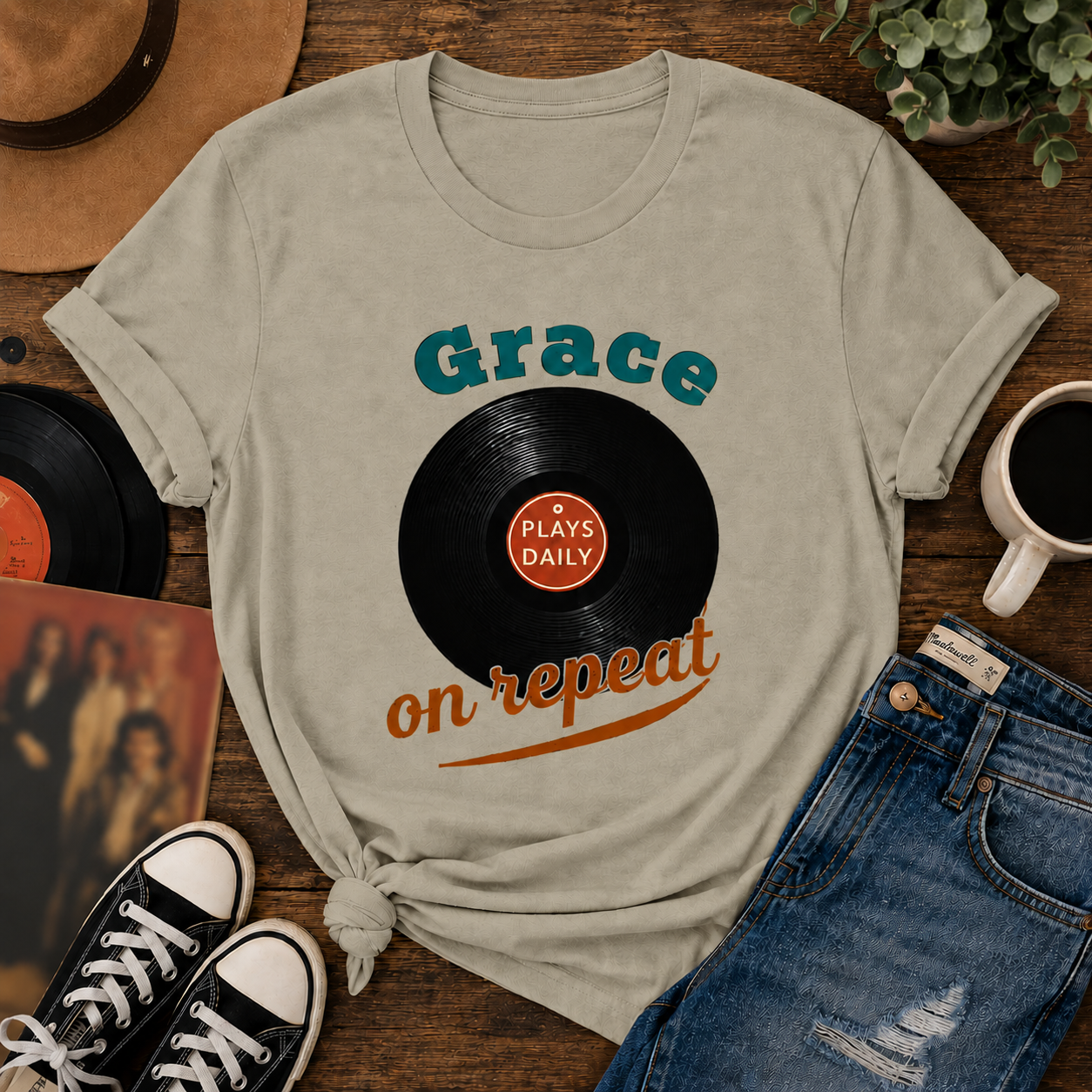 Vintage-inspired flat lay featuring a Comfort Colors Sandstone graphic t-shirt with a retro vinyl record design and the phrase “Grace Plays Daily on Repeat,” styled with denim jeans, black sneakers, coffee, and warm nostalgic Americana decor.