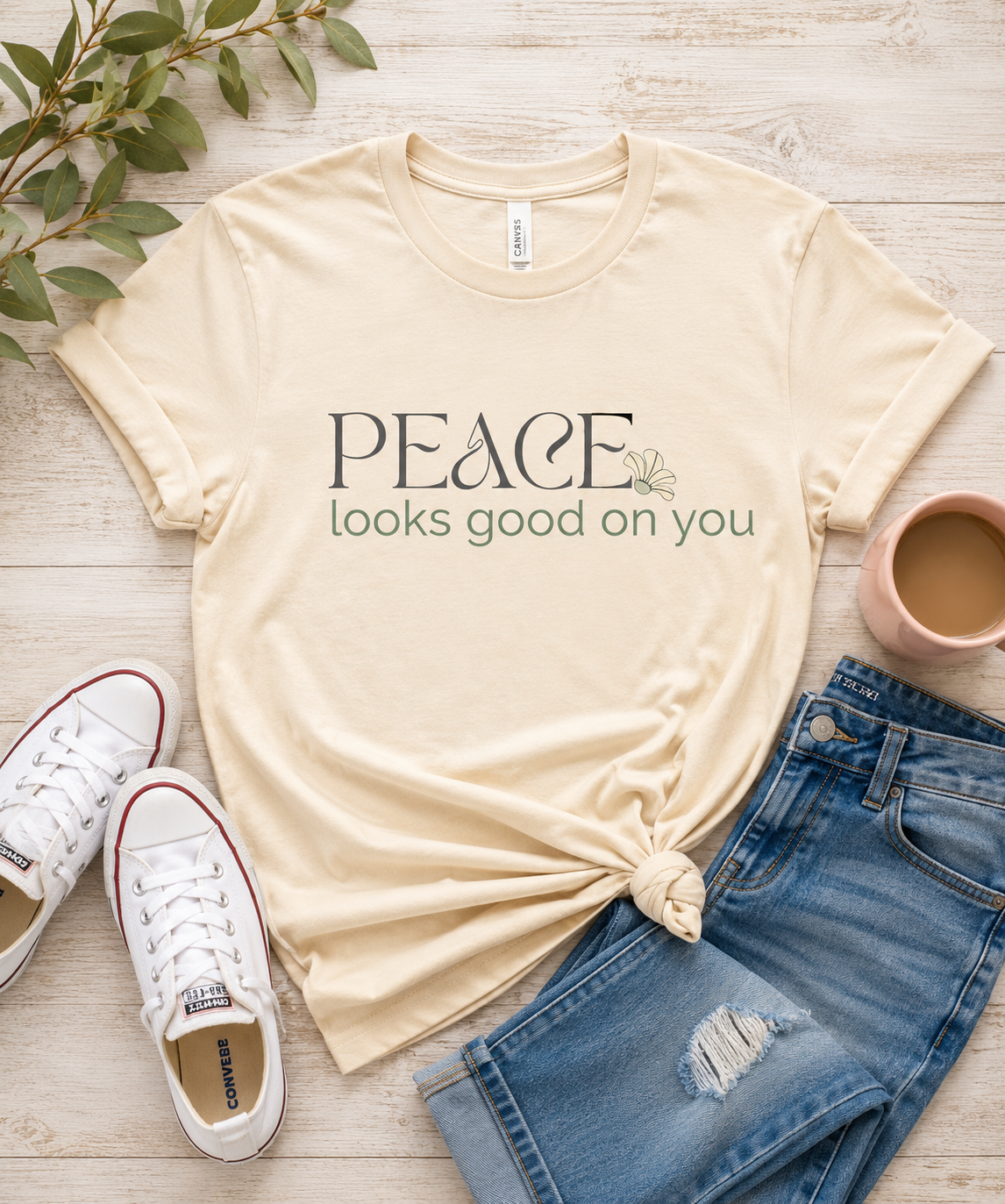 Flat lay of a cream Peace Looks Good on You graphic t-shirt styled with denim jeans, white sneakers, and a coffee mug on a light wood background.
