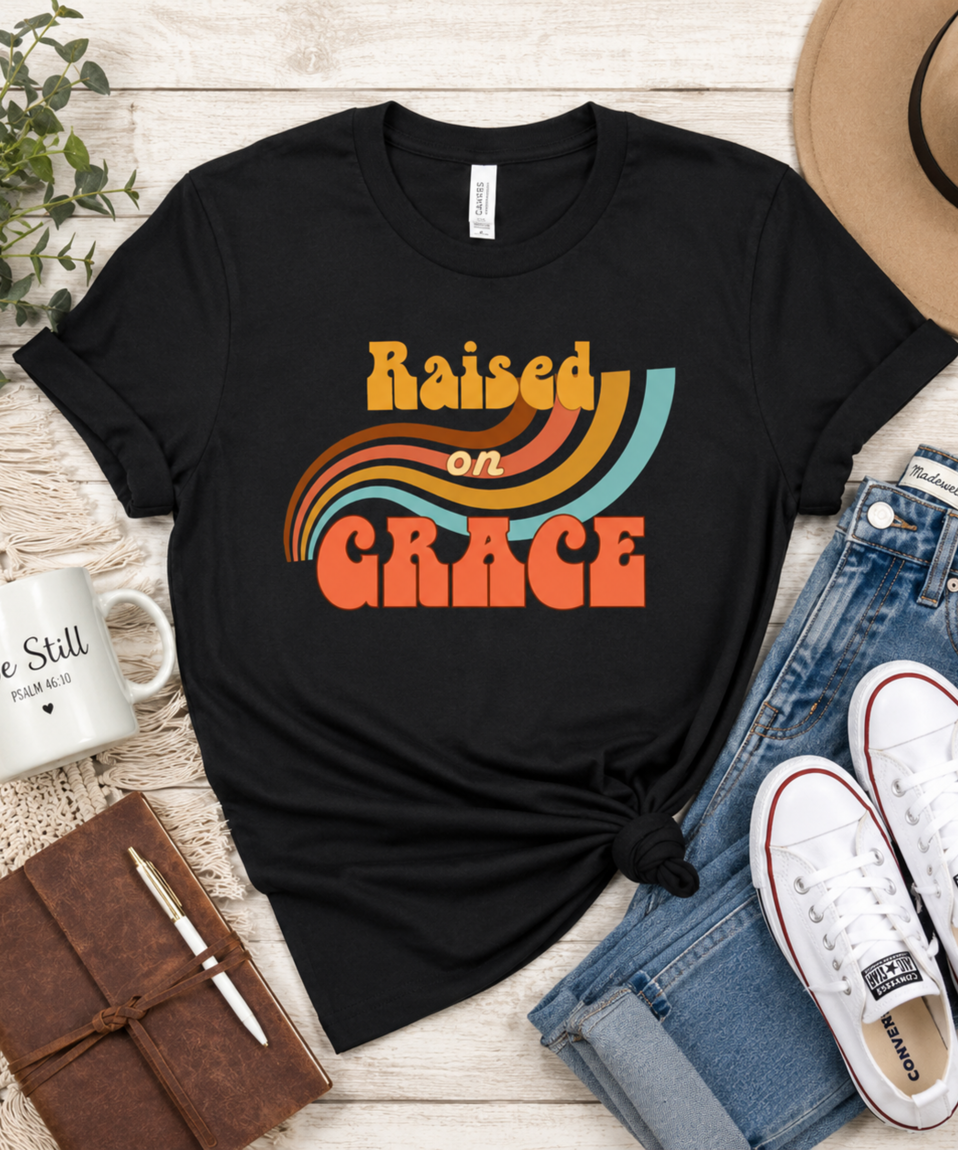 Flat lay of a black retro Christian “Raised on Grace” t-shirt styled with denim jeans, sneakers, journal, coffee mug, greenery, and neutral boho accessories on a light wood background.