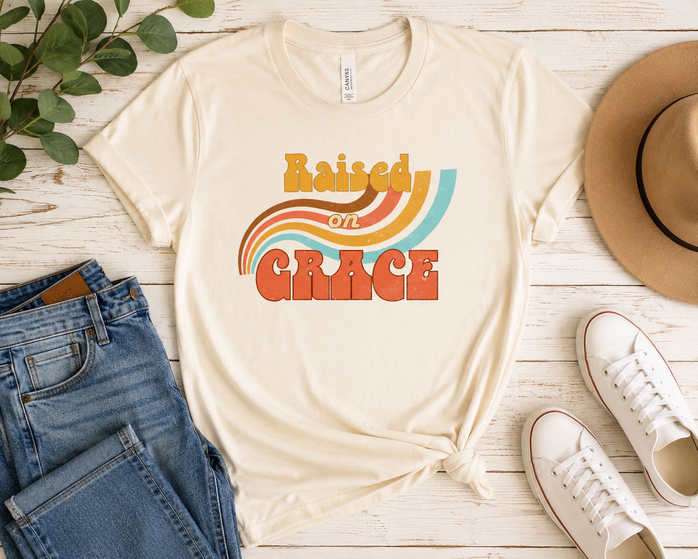 Raised on Grace distressed vintage graphic tee featuring retro typography and a worn-in nostalgic design “ t-shirt flat lay with blue jeans, tan hat, and white sneakers on a rustic white wood background.