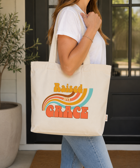 Woman carrying a natural Comfort Colors Everyday Zip Tote featuring the retro Raised on Grace design in warm mustard, coral, brown, cream, and aqua colors. Casual lifestyle mockup showcasing a vintage-inspired faith tote with a nostalgic rainbow graphic and uplifting Christian message.
