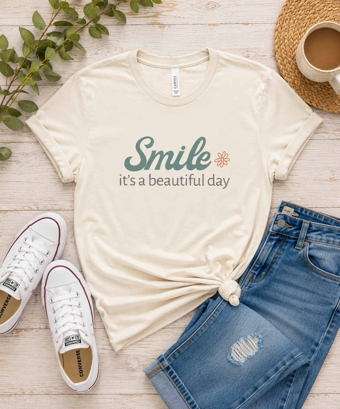 Flat lay of a cream Smile It's a Beautiful Day graphic t-shirt styled with denim jeans, white sneakers, coffee, and greenery.