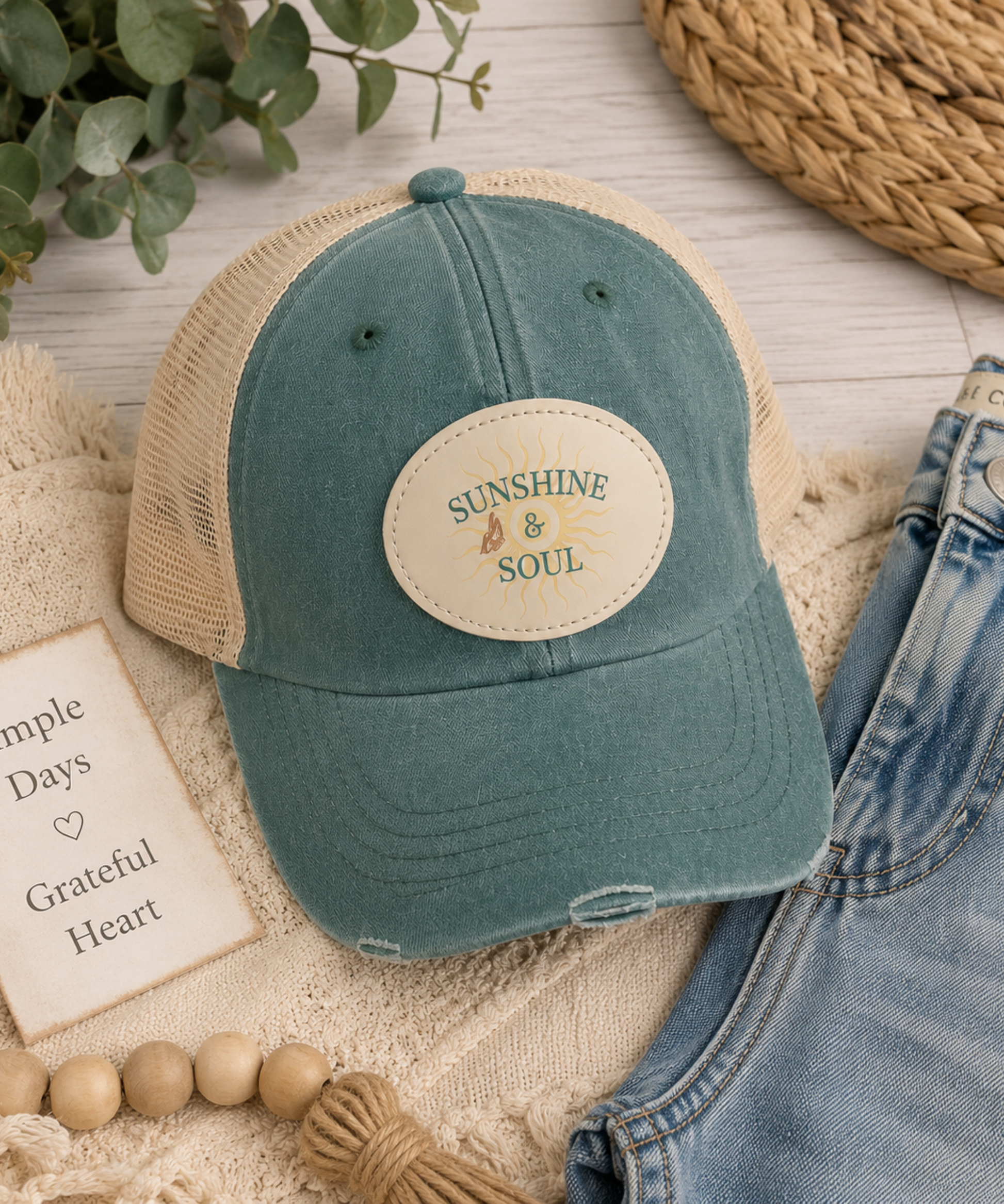 a flat lay vintage forest distressed trucker cap featuring an original Sunshine & Soul sublimated vegan leather patch with breathable mesh back and adjustable snapback closure. This cap is paired with faded jeans.